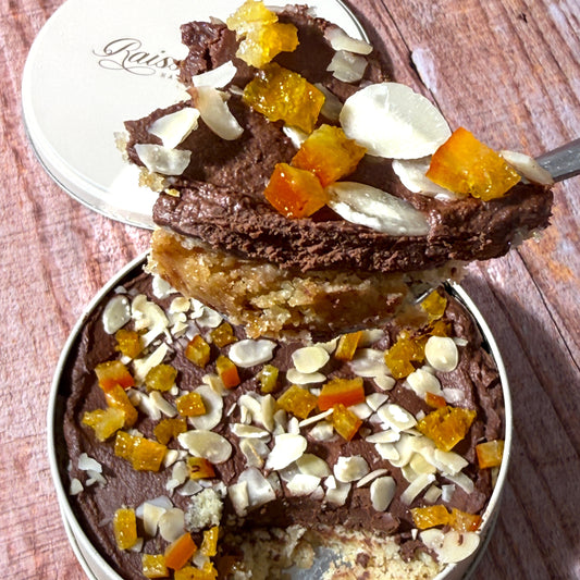 A close-up of a chocolate dessert or cake in a round metal tin, topped with sliced almonds, candied fruit pieces, and a dark chocolate glaze. A spoonful of the dessert is lifted above the tin, showing the layered interior.