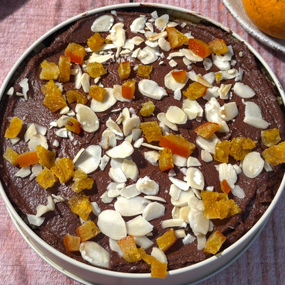 A round metal baking tin filled with a dark chocolate mousse or cake, topped with sliced almonds and candied orange fruit pieces, set on a rustic pinkish wooden surface with oranges partially visible in the background.