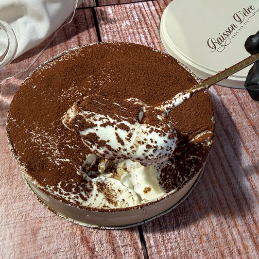 A round dessert tin filled with a chocolate-covered cream or ice cream dessert, shown on a rustic wooden table. A spoon is lifting a scoop of the dessert, and a branded lid reading 'Gaïssin Clâre' is visible in the background.