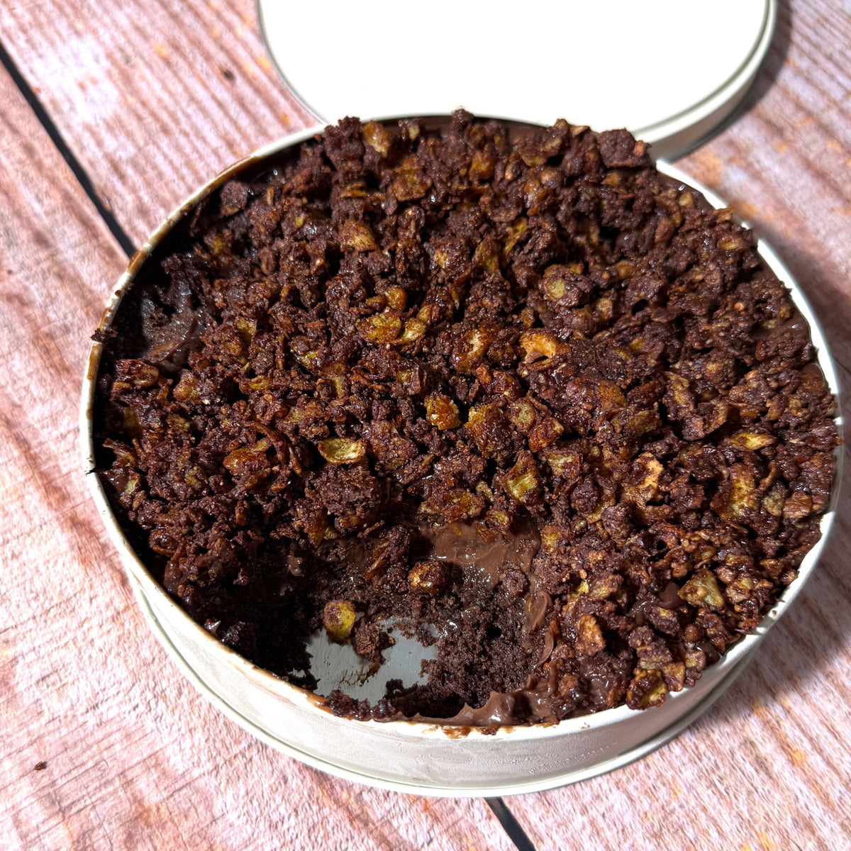 A round metal tin filled with a dark chocolate dessert or crumb cake, shown on a rustic wooden surface with the branded lid resting behind it.