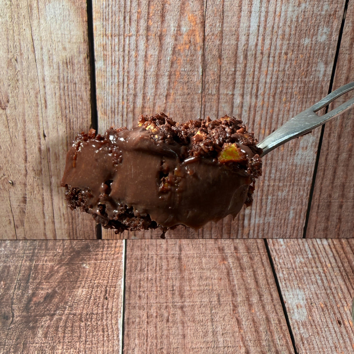 A close-up of a spoonful of chocolate cake or dessert with crumb topping, held by a hand against a rustic wooden plank background. Part of a round container lid and dark crumbs are visible in the lower right corner.