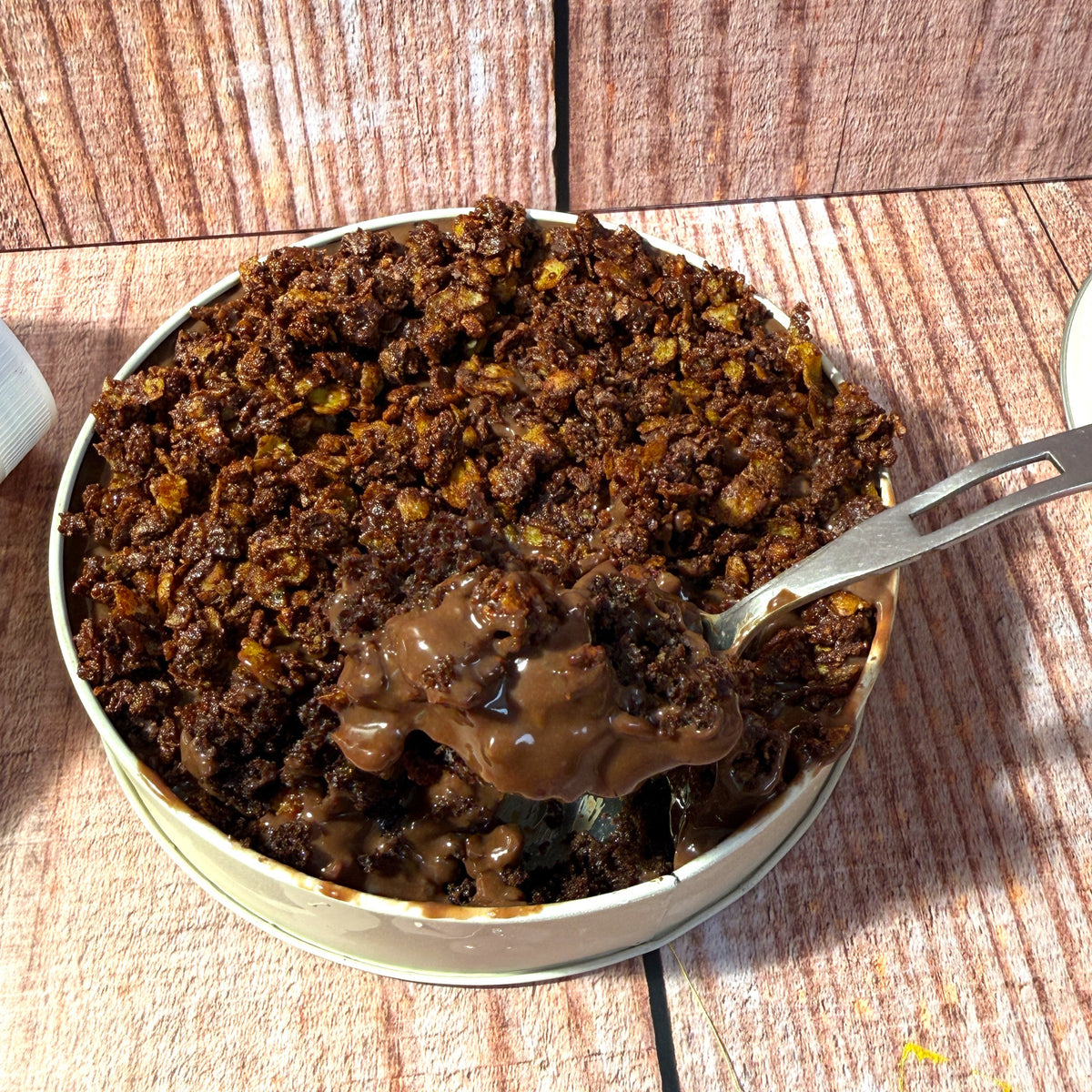 A close-up of a metal tin filled with a dark brown crumbly dessert or chocolate mixture, with a spoon lifting a scoop. A hand is holding the spoon on a rustic wood plank surface.