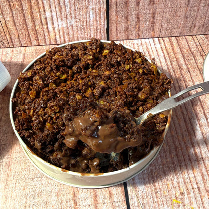A close-up of a metal tin filled with a dark brown crumbly dessert or chocolate mixture, with a spoon lifting a scoop. A hand is holding the spoon on a rustic wood plank surface.